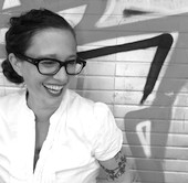 Das Portrait zeigt Simone Veenstra. Sie lacht, trägt eine dunkle Brille und ihre Haare sind zu einem tiefen Dutt gebunden. Im Hintergrund ist ein Stück Graffiti und eine Mauer zu sehen.